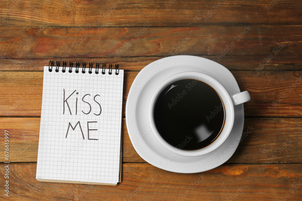 Notebook with phrase Kiss Me and cup of coffee on wooden table, flat lay Notebook with phrase Kiss Me and cup of coffee on wooden table, flat lay