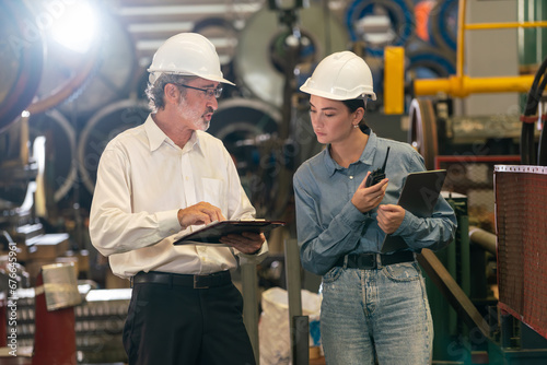 Factory engineer manager with assistant using laptop to conduct inspection of steel industrial machine, exemplifying leadership as machinery engineering inspection supervisor in metalwork manufacture.