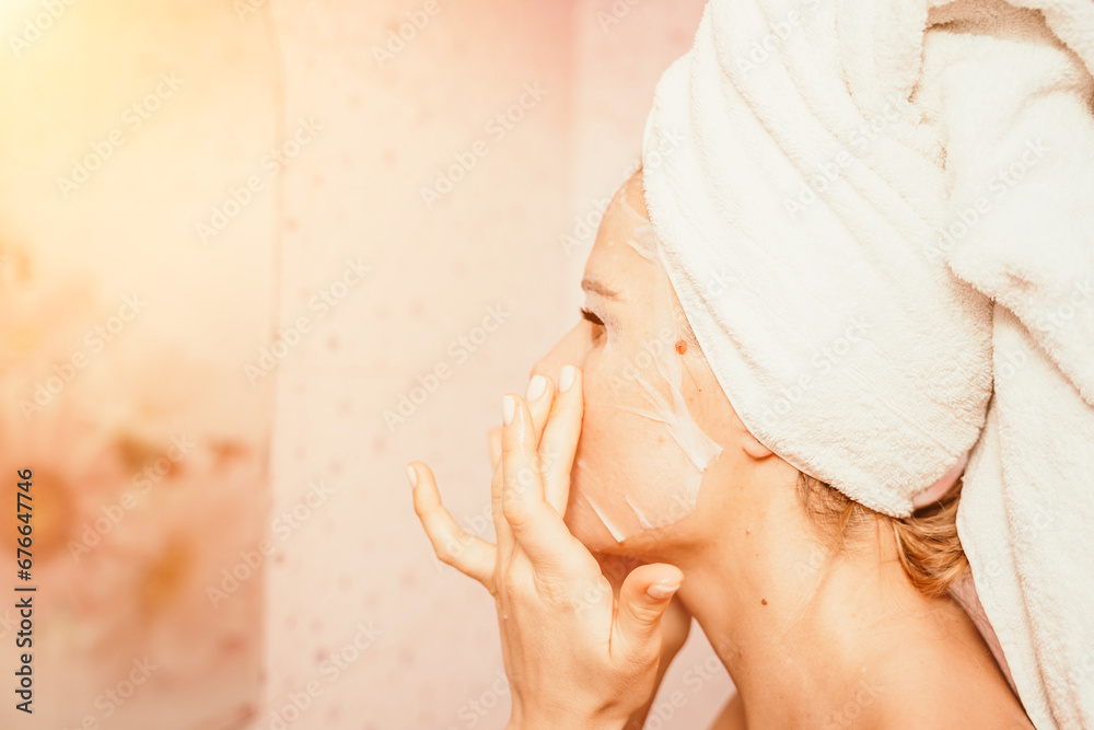 Naklejka premium Young beautiful woman using a moisturizing facial mask after taking a bath. Pretty attractive girl in a towel on her head stands in front of a mirror in a home bathroom. Daily hygiene and skin care