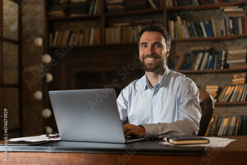 A smiling man sits at a table in the library and uses a laptop. A young scientist prepares to defend candidate or doctoral dissertation. A joyful businessman receives an offer for profitable