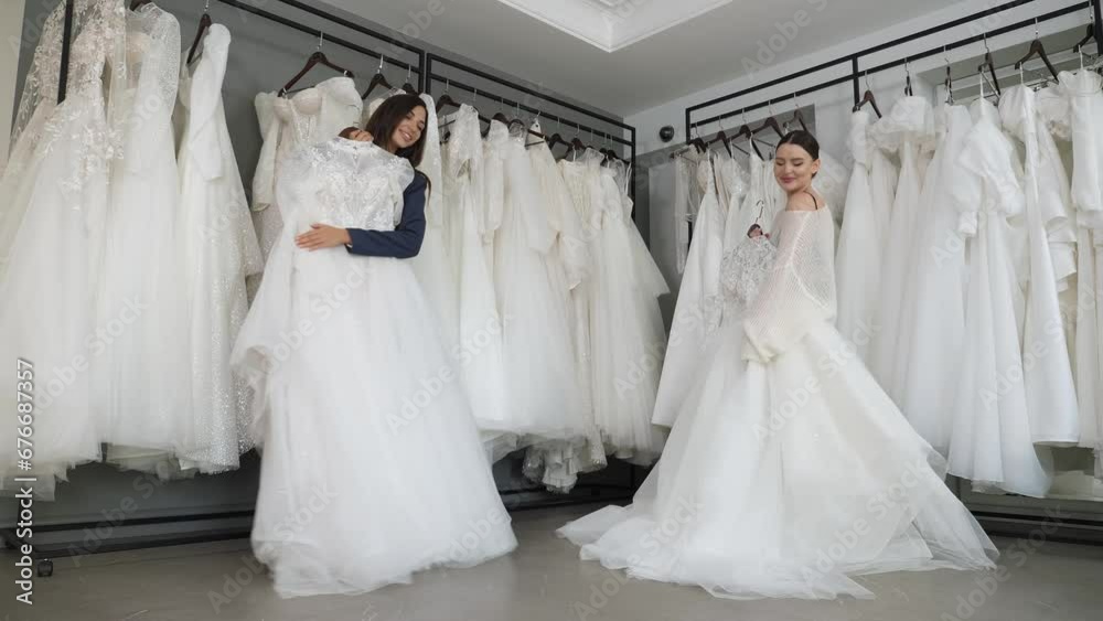 Portrait of two happy young brides in a wedding salon, they are holding wedding dresses in their hands and dancing. Girlfriends choose a wedding dress together. Preparation for the wedding.