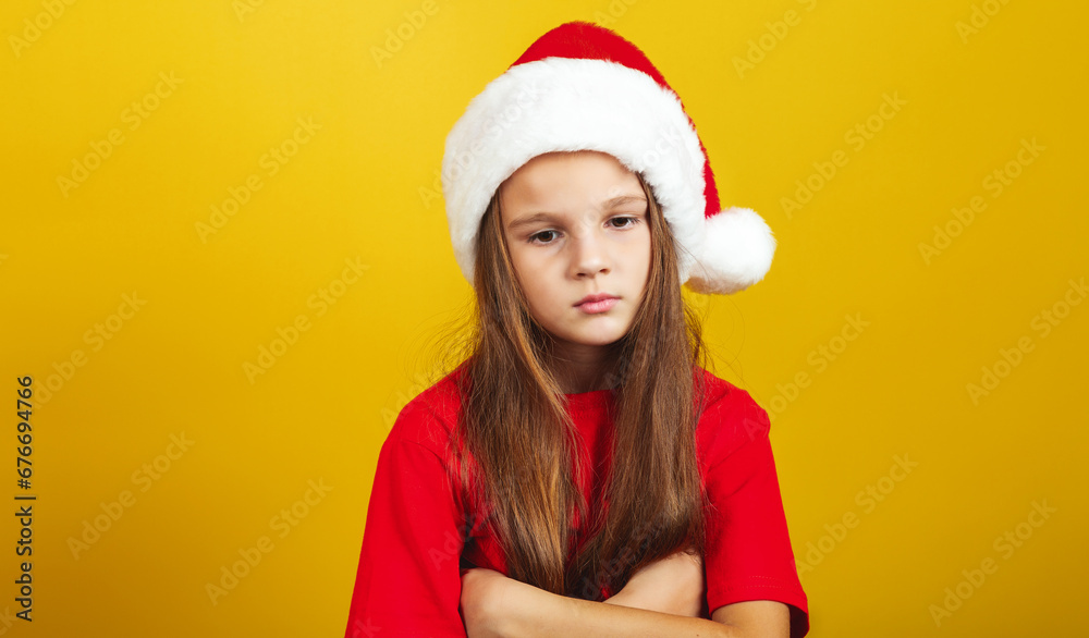 Pensive little girl wearing Christmas costume and Santa hat standing isolated over yellow background wall in studio Pensive little girl wearing Christmas costume and Santa hat standing isolated over yellow background wall in studio