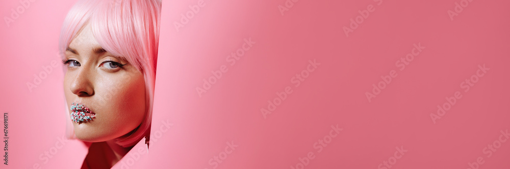 Face of a young beautiful girl model with bright makeup and lips with glitter looks into a hole in pink paper. Beauty, fashion, personal care, cosmetics, makeup, beauty salon. Banner with copy space. - obrazy, fototapety, plakaty 