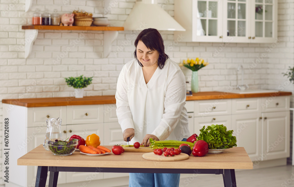Cheerful overweight woman preparing proper meal in kitchen. Smiling plus size, plump young woman cutting vegetables at kitchen table at home. Healthy food, dieting concept
