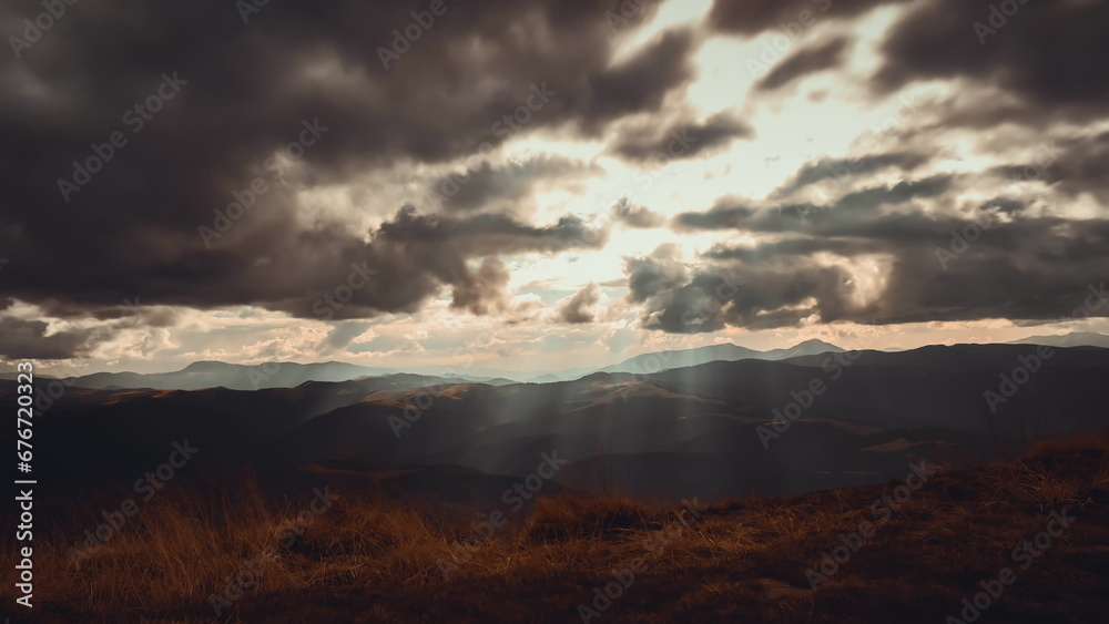 Dark dramatic autumn mountain cloudscape. Epic storm clouds moving fast, rays of rising sun break through the clouds hit the orange meadow. Vintage dark sunset. Nature background