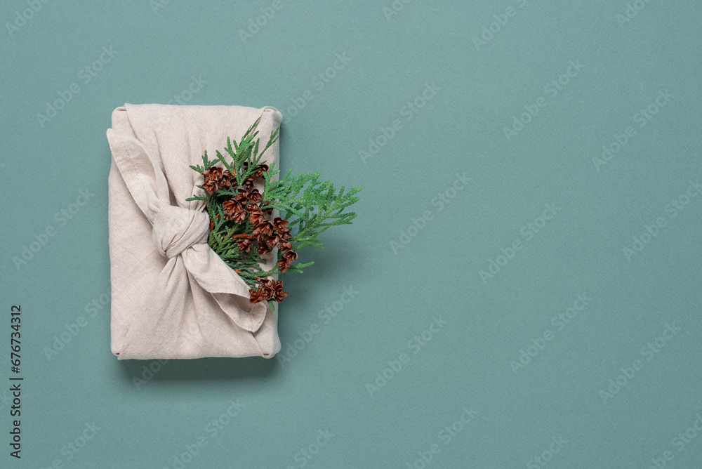 Christmas gift wrapped in fabric with conifer branch, green pastel background. A traditional furoshiki gift. Top view,flat lay. - obrazy, fototapety, plakaty 