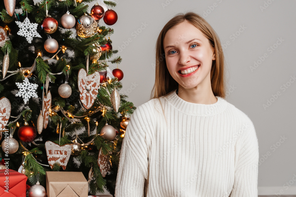 the girl smiles at the camera next to the Christmas tree. A blonde girl in a white sweater stands in a bright room with a decorated Christmas tree in the background. festive mood concept