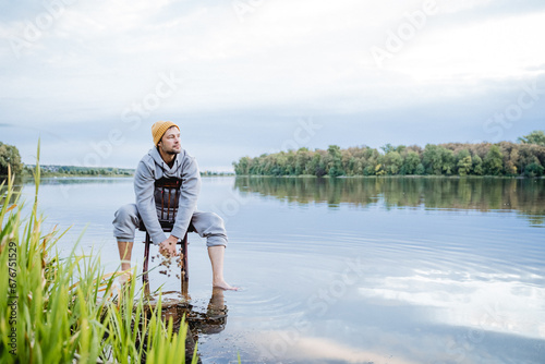 A man is sitting in the water on an old chair and looking at the lake, a guy is admiring the beautiful nature, a hipster is standing barefoot in the water.
