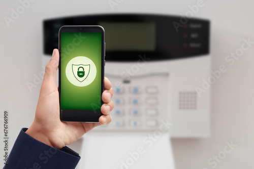 Man operating home alarm system via mobile phone against white wall with security control panel, closeup. Application with illustration of padlock in shield on device screen