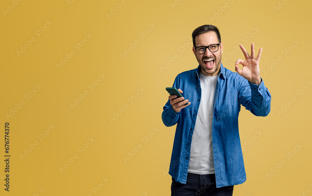 Handsome freelancer shouting happily and showing OK sign while using cellphone on yellow background Handsome freelancer shouting happily and showing OK sign while using cellphone on yellow background