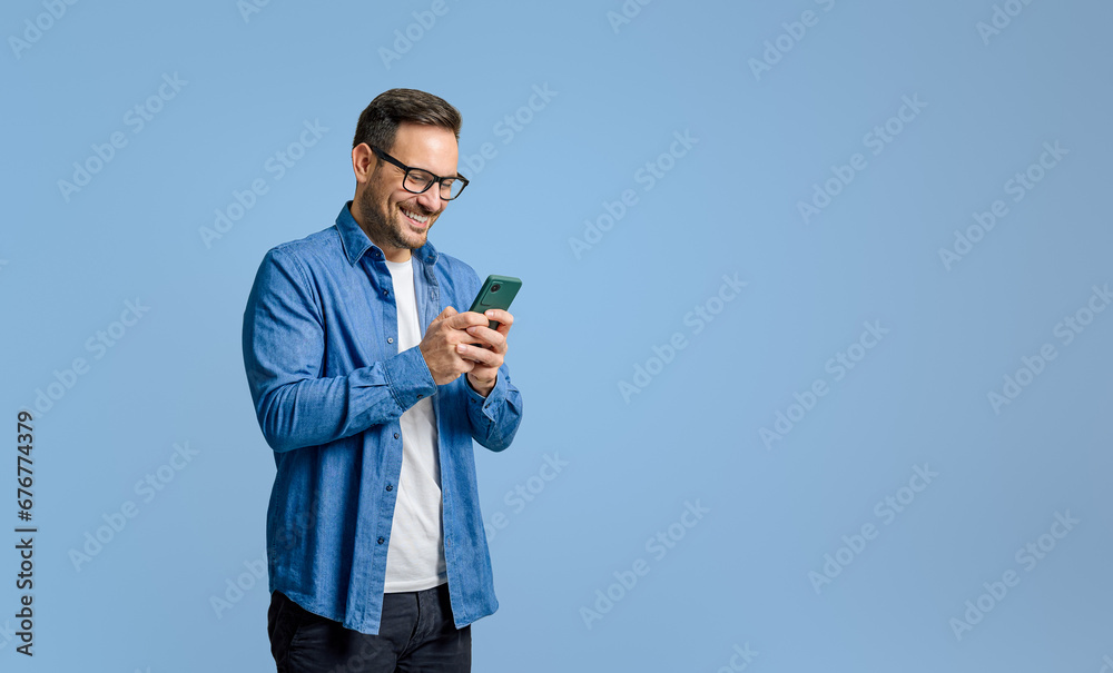 © Moon Safari - Smiling handsome businessman text messaging over mobile phone while standing on blue background © Moon Safari - Smiling handsome businessman text messaging over mobile phone while standing on blue background