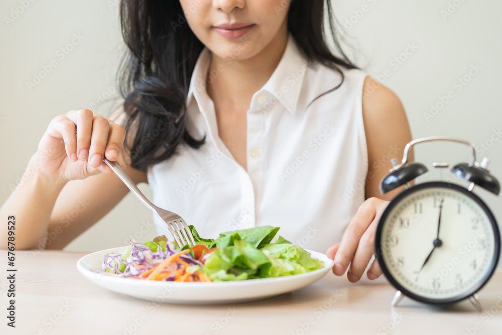 Intermittent fasting with clock, health asian young woman, girl weight loss, eating green fresh vegetable salad on dish, plate with eat healthy of breakfast food in morning, lunch on a table at home. Intermittent fasting with clock, health asian young woman, girl weight loss, eating green fresh vegetable salad on dish, plate with eat healthy of breakfast food in morning, lunch on a table at home.
