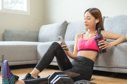 Diet meal replacement for weight loss, asian young woman sitting on floor in sportswear, drinking protein shake, having sports supplement for muscle, using smart phone, earphone after workout at home.