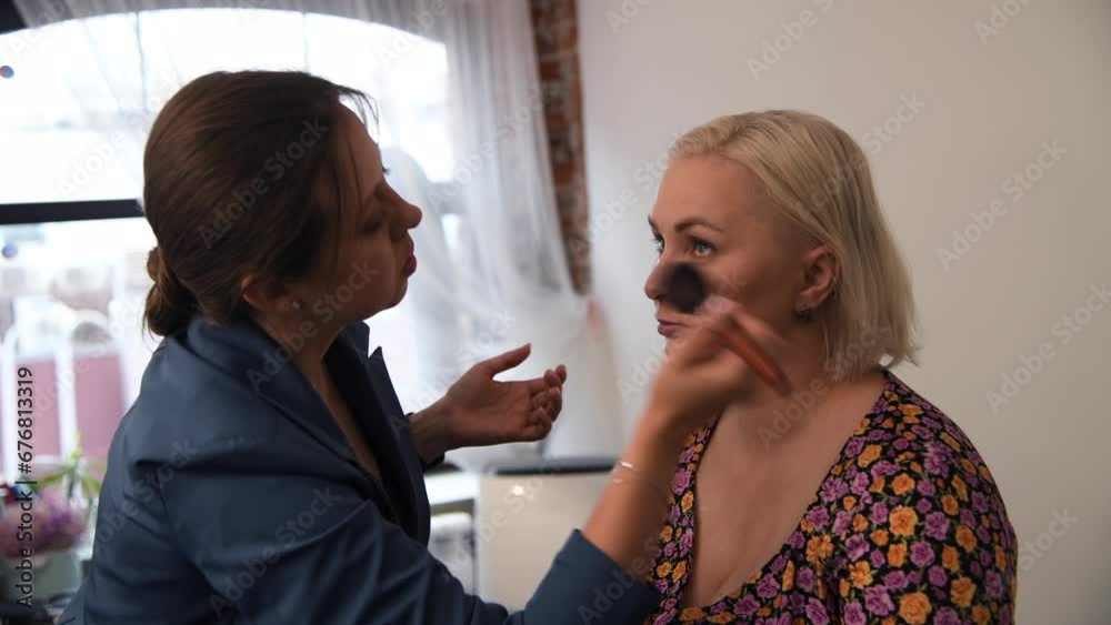 Side view of make-up artist applying powder makeup foundation using brush on adult blond woman's skin near eyes in beauty salon. Soft focus. Real time handheld video. Make-up theme. Side view of make-up artist applying powder makeup foundation using brush on adult blond woman's skin near eyes in beauty salon. Soft focus. Real time handheld video. Make-up theme.