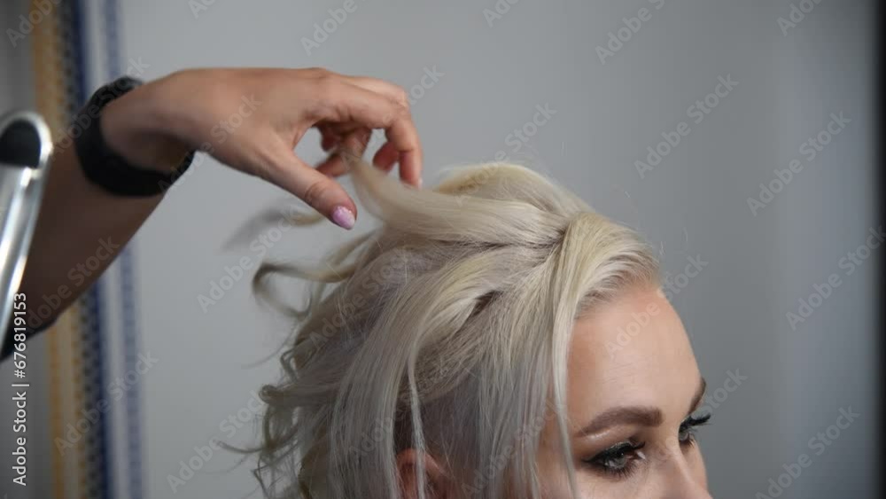 Close-up view of hairdresser making new coiffure for adult blond haired woman using curling iron and comb in hair salon. Soft focus. Real time handheld video. Beauty industry theme.