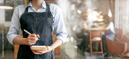 Female waitress wearing apron standing in cozy coffeehouse with notepad in hands to take order in restaurant photo
