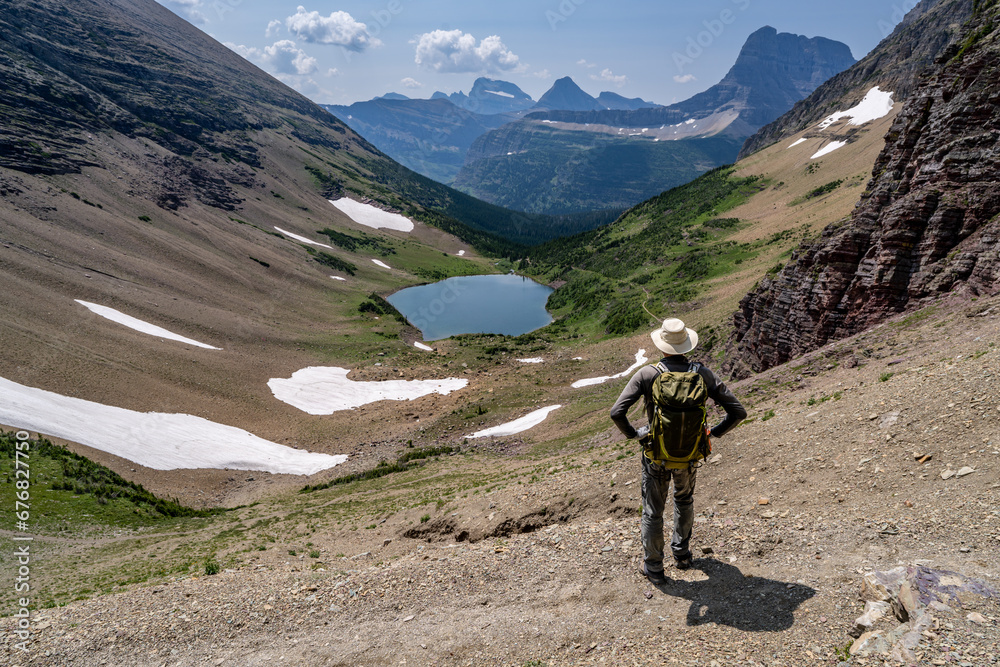 Mature Caucasian Man Hiker standing watching an alpine scene of a lake in a deep canyon surrounded by mountains, Ptarmigan Lake, Glacier National Park, Montana Mature Caucasian Man Hiker standing watching an alpine scene of a lake in a deep canyon surrounded by mountains, Ptarmigan Lake, Glacier National Park, Montana