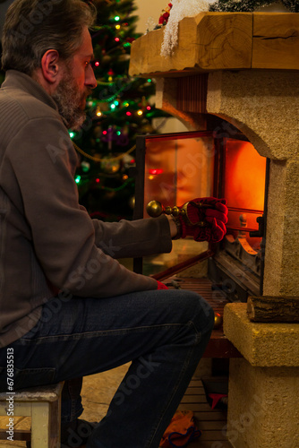 Man sits in front of burning fireplace. Behind Christmas tree