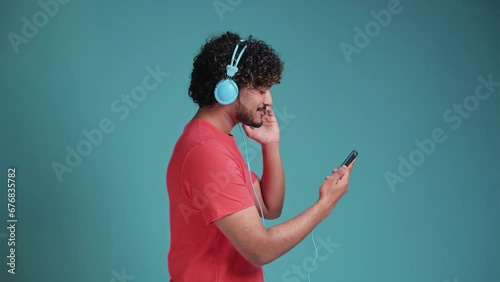 handsome young indian guy in casual listen music on smartphone, using new headset, looking at cell phone and smiling, in coral t-shirt on blue studio background