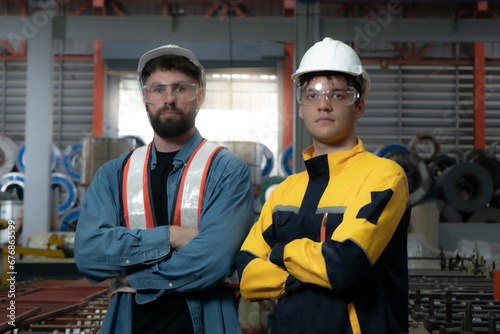 A man in a blue shirt and safety vest stands in front of a stack of metal pipes. He is wearing a hard hat and he is a worker in a factory