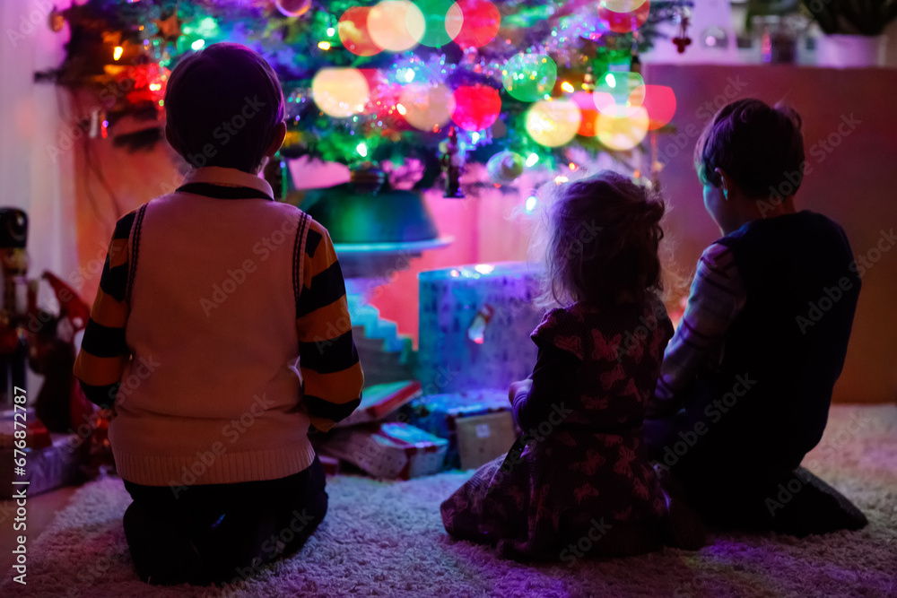Two little kid boys and adorable toddler girl sitting by Christmas tree with old vintage toys and balls with lots of present on Xmas eve. Family preaparation celebration of family feast