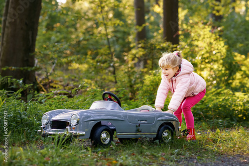 Little preschool girl driving big vintage toy car. Happy child having fun with playing outdoors. Active preschooler child enjoying warm autumn day in forest. Smiling stunning kid playing © Irina Schmidt