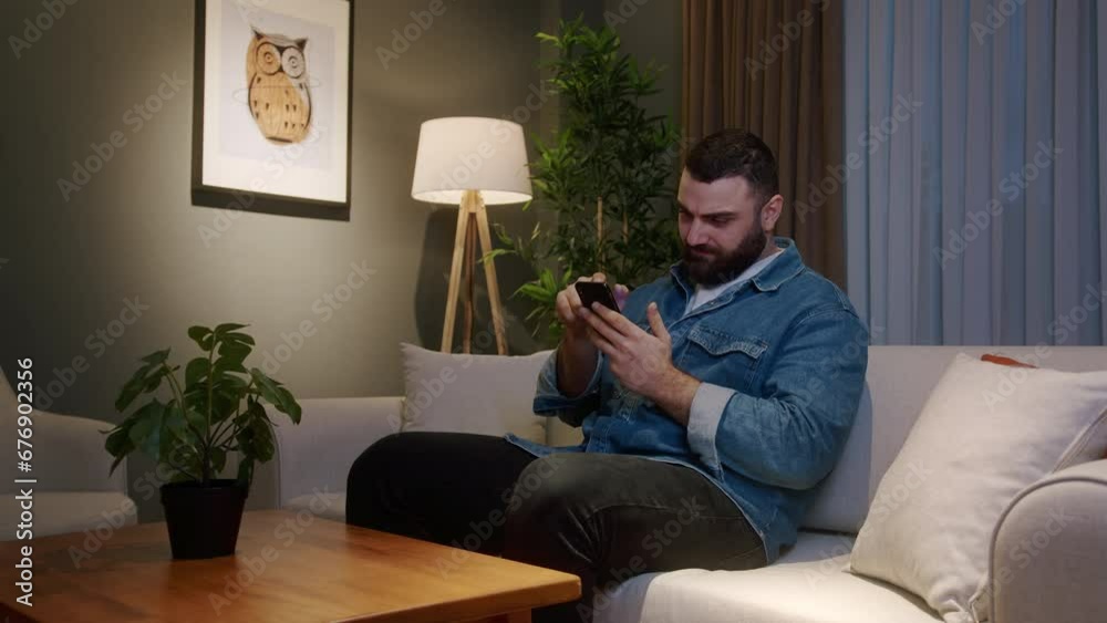 Handsome bearded young adult man sitting and relaxing on couch at home holding a smartphone enjoying surfing internet, using social media or app. Modern tech device at home
Handsome bearded young adult man sitting and relaxing on couch at home holding a smartphone enjoying surfing internet, using social media or app. Modern tech device at home