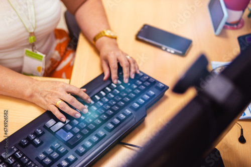 Senior woman working on a computer, her hands in the foreground; the PC is black in the office. There is a blue glow, and a cell phone is placed next to the keyboard.