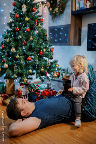 Little smiling girl with a reflex camera in her hands sits on her dad stomach and looks at him near the Christmas tree