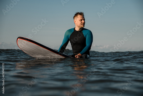 Young male surfer sitting on a surfboard and sea looking thoughtfully into the distance against a background of blue sky