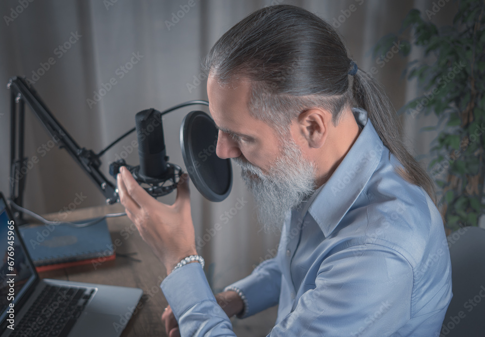 
A man with a beard wearing glasses broadcasts his audio podcast using a microphone and laptop in a home studio. Close-up
