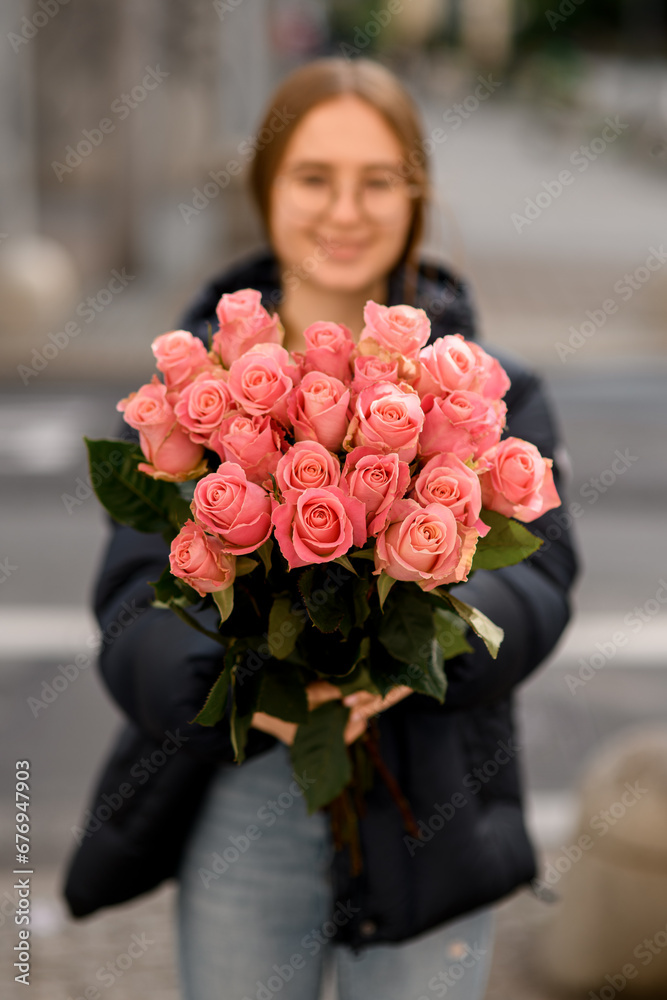 Fototapeta premium Young pretty dark-haired girl stands on a city street with a bouquet of pink roses