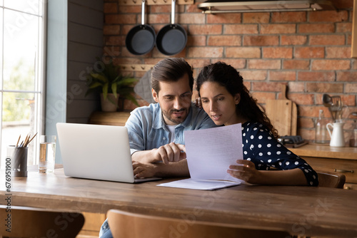 Young couple checking financial documents together, discussing loan agreement, sitting at table with laptop in kitchen, wife and husband calculating taxes, domestic bills, family planning budget photo