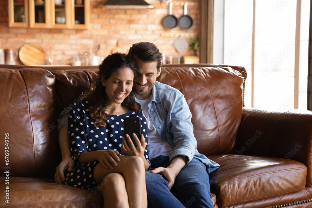 Happy young couple hugging, having fun with smartphone together, relaxing sitting on cozy couch, smiling excited woman and man looking at phone screen, making video call, chatting or shopping online Happy young couple hugging, having fun with smartphone together, relaxing sitting on cozy couch, smiling excited woman and man looking at phone screen, making video call, chatting or shopping online