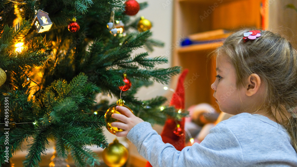 A little charming girl in a dress hangs a shiny ball on the Christmas tree. A child is decorating a Christmas tree in a beautiful living room. The concept of a happy family holiday New Year A little charming girl in a dress hangs a shiny ball on the Christmas tree. A child is decorating a Christmas tree in a beautiful living room. The concept of a happy family holiday New Year