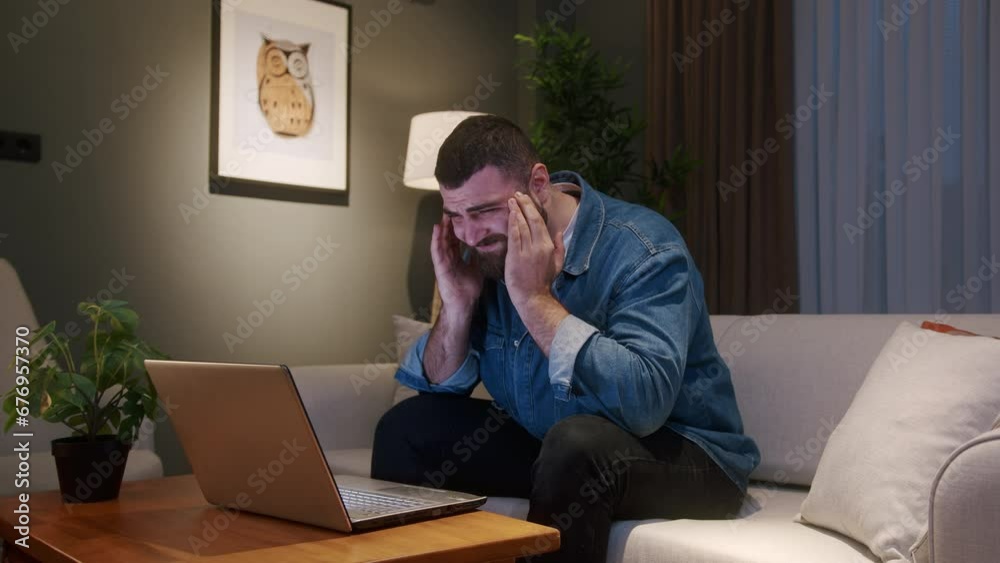 Young adult man sitting on sofa at home while using laptop having headache. Tired young man get having pain from a headache or migraine Young adult man sitting on sofa at home while using laptop having headache. Tired young man get having pain from a headache or migraine