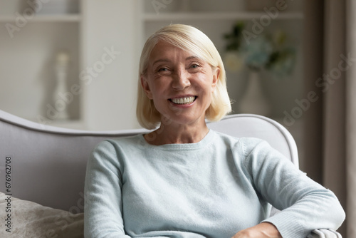 Head shot portrait mature woman with healthy toothy smile, beautiful older female with grey hair looking at camera, sitting on cozy sofa at home, posing for photo, making video call, using webcam