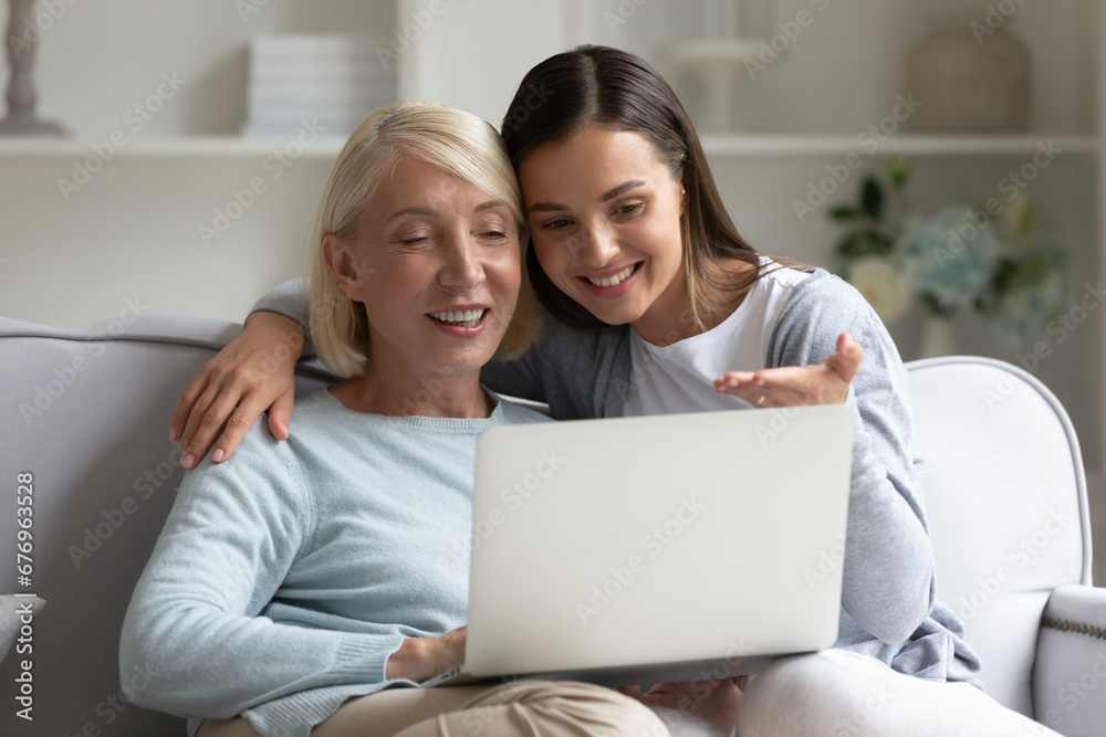 Happy mature mother and adult daughter using laptop, surfing internet or shopping online, older grandmother and granddaughter looking at computer screen, watching video or movie, having fun at home Happy mature mother and adult daughter using laptop, surfing internet or shopping online, older grandmother and granddaughter looking at computer screen, watching video or movie, having fun at home