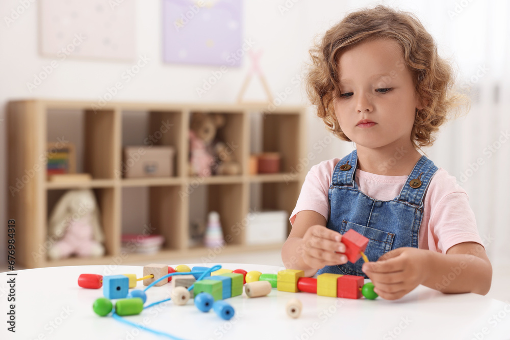 Motor skills development. Little girl playing with wooden pieces and string for threading activity at table indoors Motor skills development. Little girl playing with wooden pieces and string for threading activity at table indoors