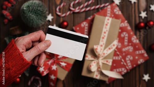 Hands of a woman in a red sweater with a credit card on a background of gift boxes, selective focus, close-up. Christmas and New Year shopping online, payment by credit card. Buying gifts online.