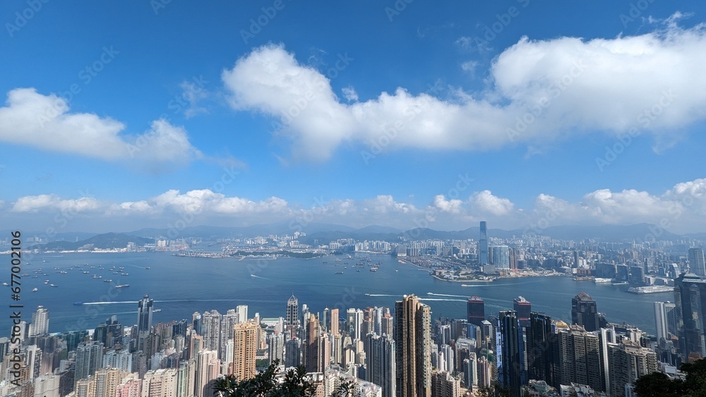 Obraz premium Panoramic Aerial View of Hong Kong Skyline and Victoria Harbour with Dense Skyscrapers, Boats, and Mountains on a Sunny Day with Blue Sky and Fluffy Clouds