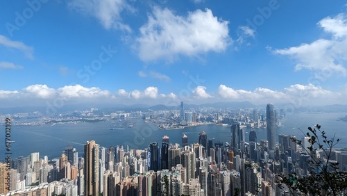 Panoramic Aerial View of Hong Kong Skyline and Victoria Harbour with Dense Skyscrapers, Boats, and Mountains on a Sunny Day with Blue Sky and Fluffy Clouds