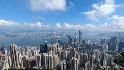 Panoramic Aerial View of Hong Kong Skyline and Victoria Harbour with Dense Skyscrapers, Boats, and Mountains on a Sunny Day with Blue Sky and Fluffy Clouds
