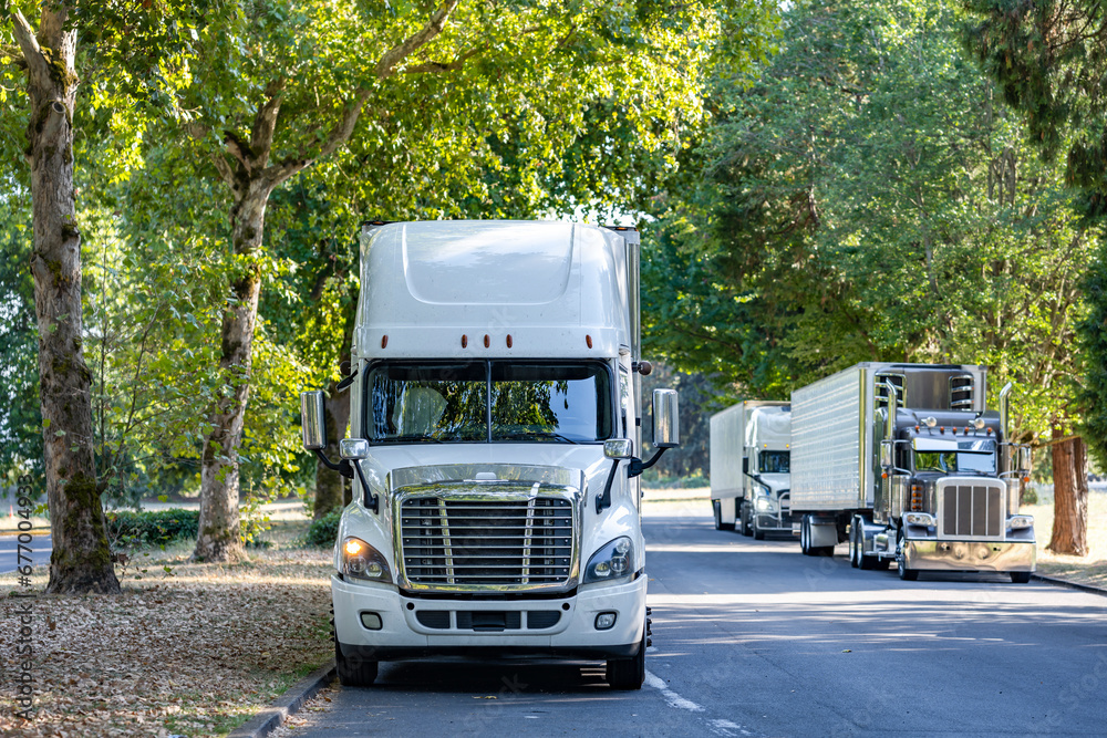 Line of the big rig semi trucks with semi trailers stand on the rest area on a shady road under spreading trees for the truck driver rest