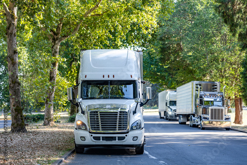Line of the big rig semi trucks with semi trailers stand on the rest area on a shady road under spreading trees for the truck driver rest
