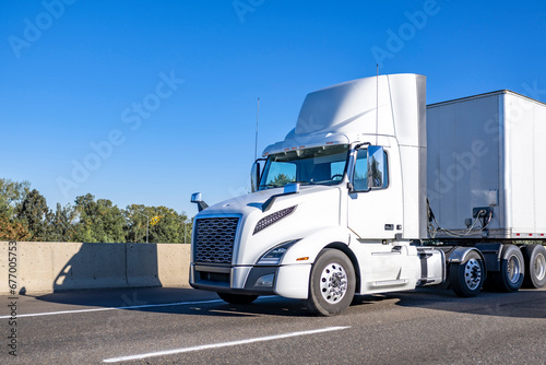 Powerful day cab white big rig semi truck with built-in roof spoiler transporting cargo in dry van semi trailer running on the road at sunny day