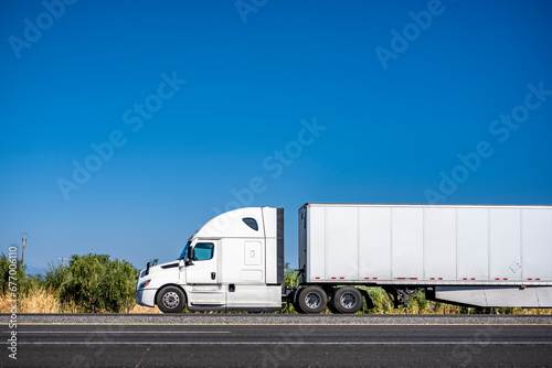 White big rig bonnet semi truck transporting commercial cargo in dry van semi trailer driving on the flat highway road