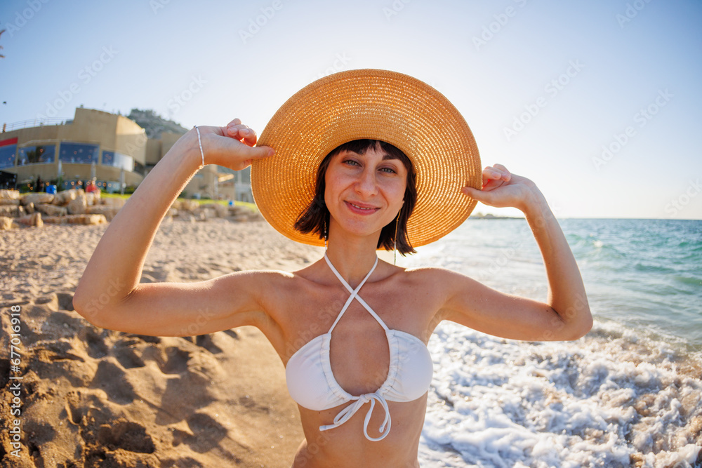 Portrait of a beautiful young woman in a straw hat on the seashore. Cheerful young woman smiling on the beach during summer. Happy girl with black hair enjoying the sun.
