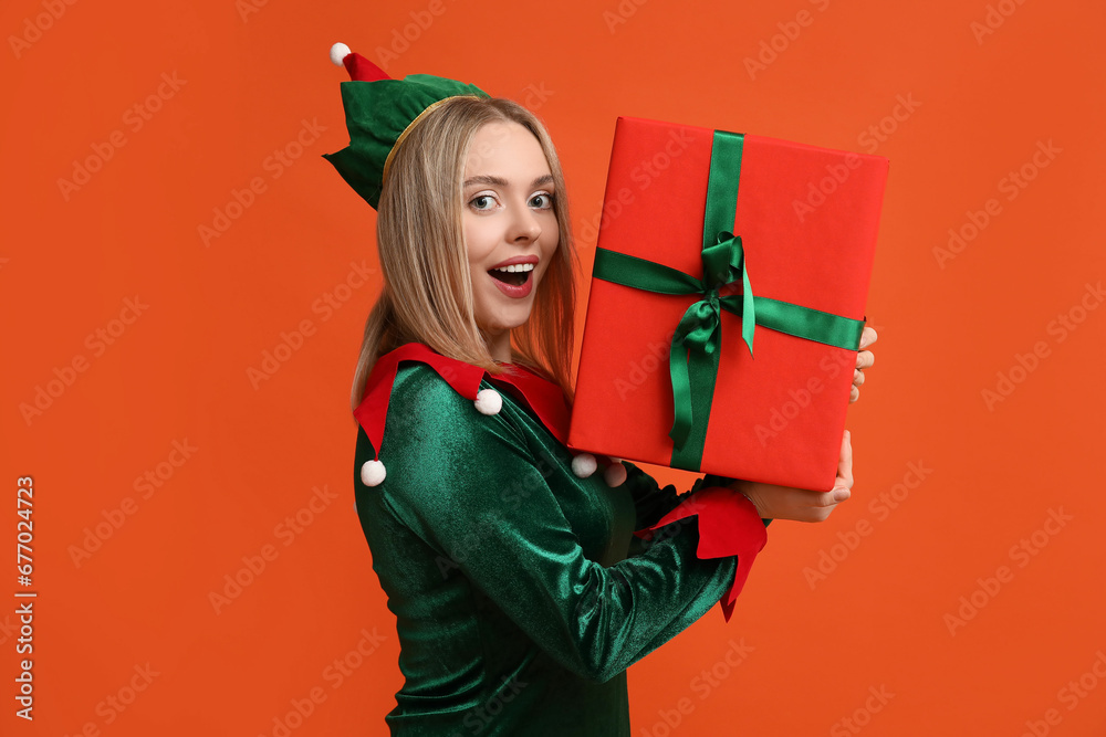Young woman in elf costume with Christmas gift box on orange background - obrazy, fototapety, plakaty 