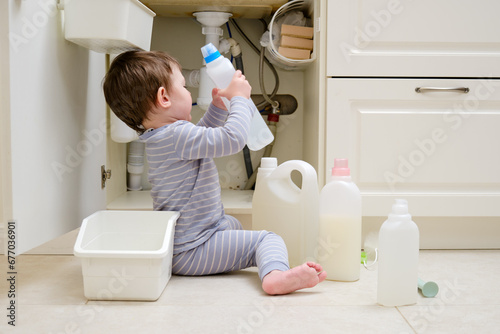A child is playing with chemical cleaning products under the sink in the kitchen. Baby holds bottles with detergent. Kid aged about two years (one year nine months) photo